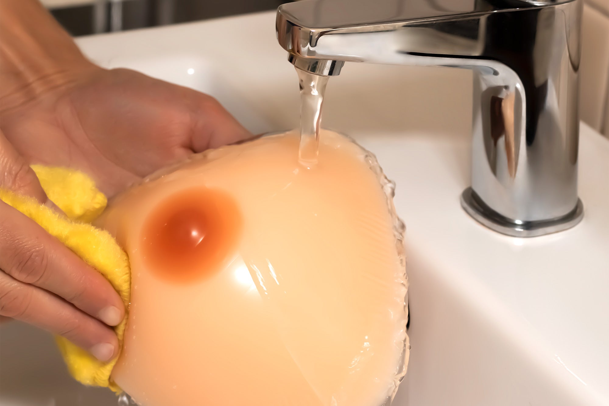 A peach-colored silicone breast form being rinsed under running water in a sink for breast form care guide