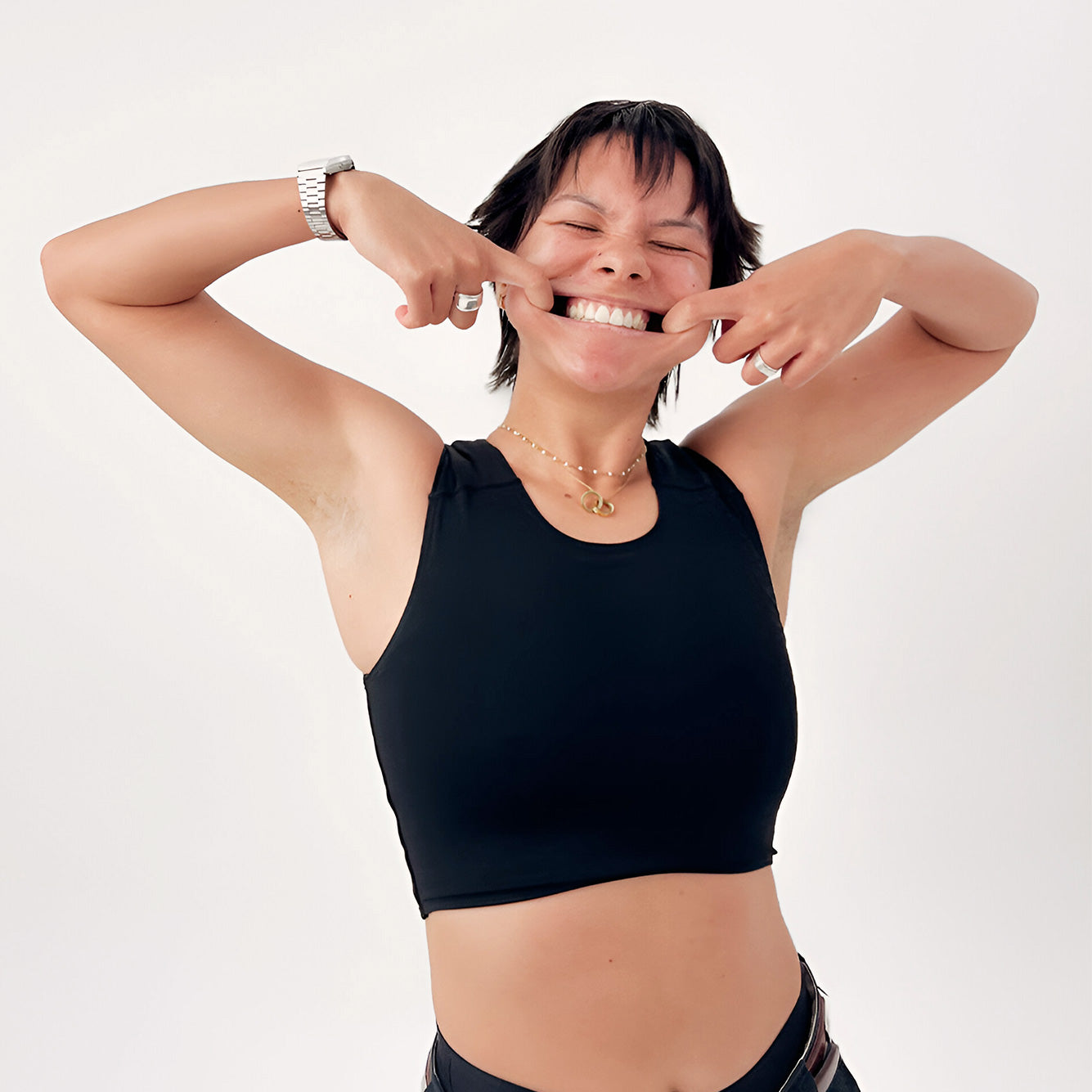 A person with short dark hair wearing a black chrest binder and black bottoms, posing with their hands pulling a smile against a plain light gray background.