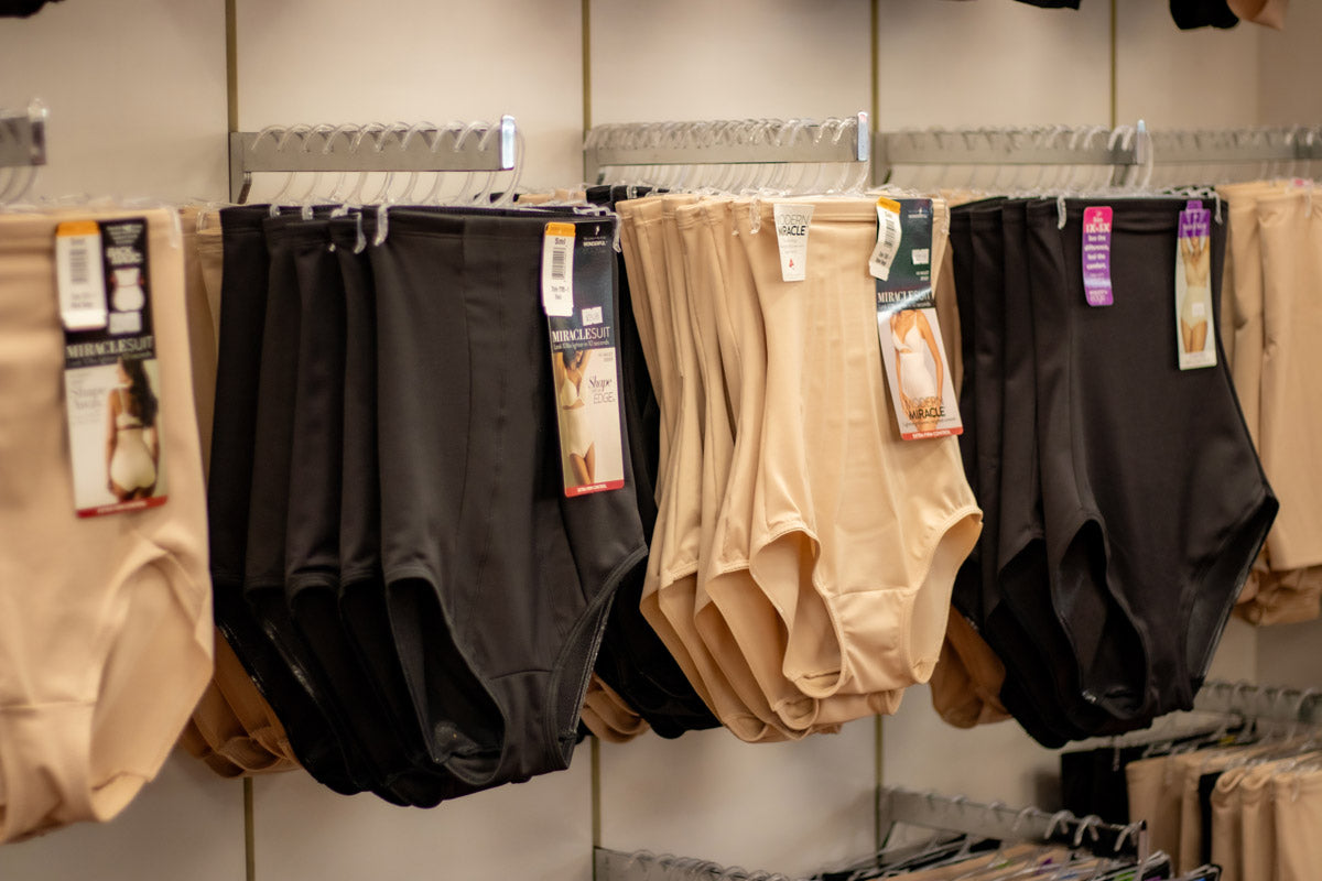 Multiple pairs of seamless high-waisted briefs for crossdressing in black and beige shades hanging on metal racks in a retail store.