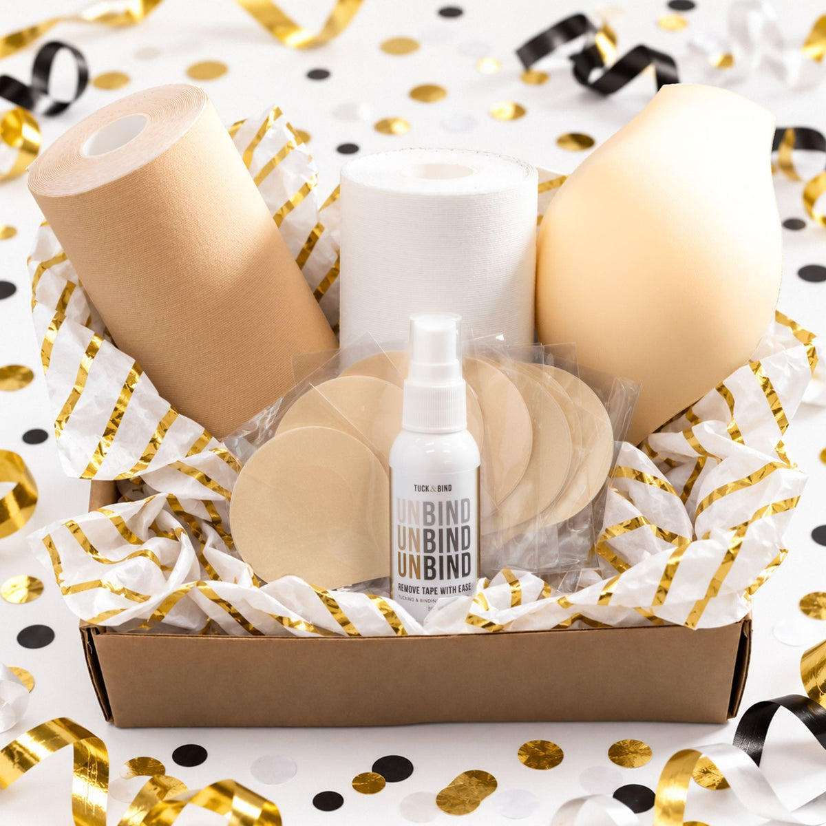 A gift box of trans masc accessories including trans tape, removal spray, nipple covers and a packer displayed on a white background with gold and black confetti.