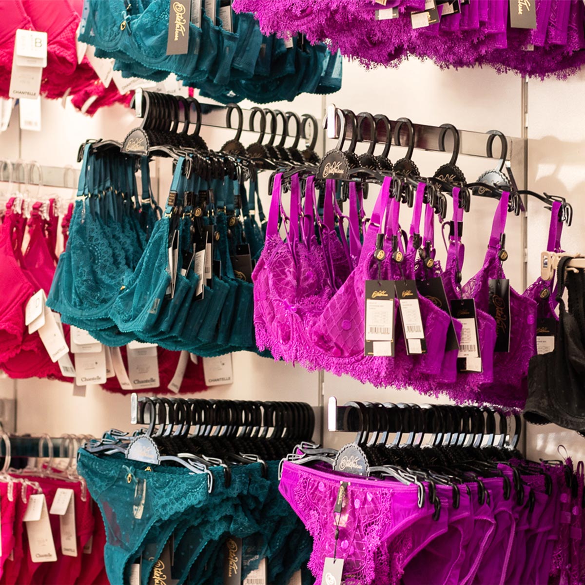 A retail display of lingerie on black hangers, featuring bright magenta lace bras and panties alongside teal lace and satin briefs and garter sets, with product tags hanging from each item.