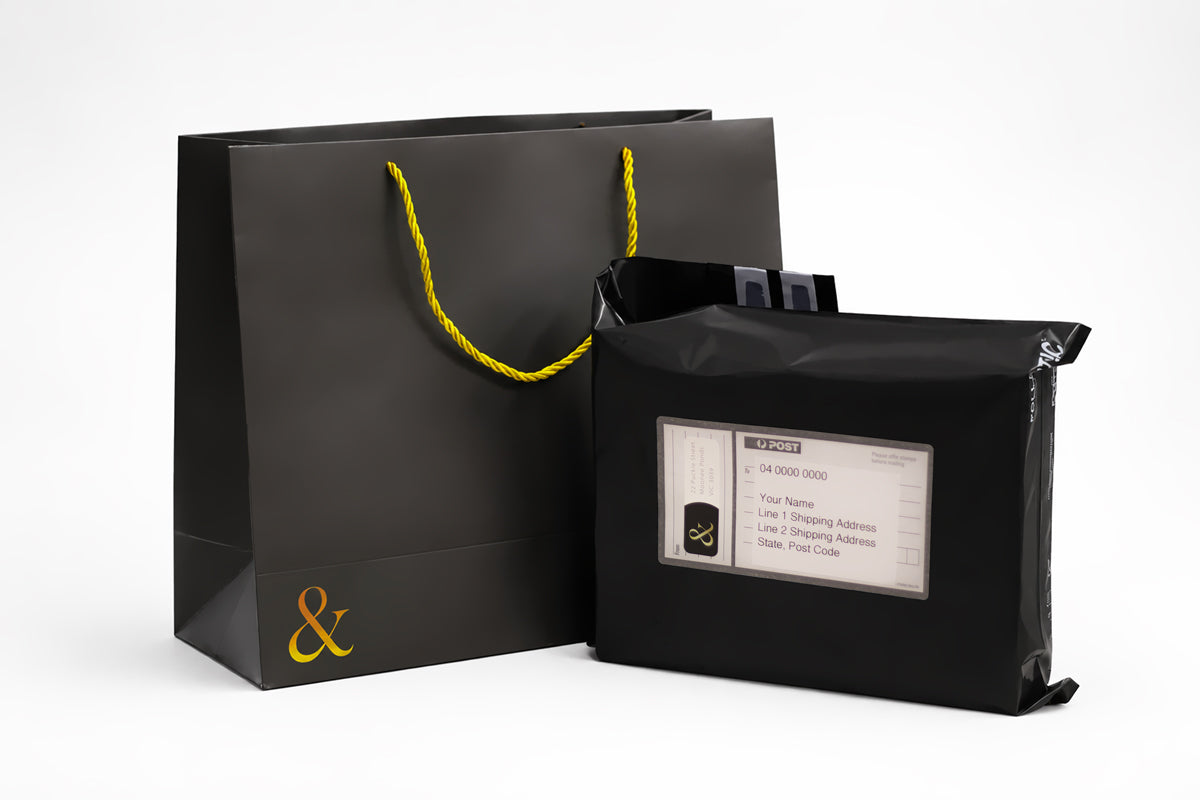Tuck & Bind black paper shopping bag with gold-colored twisted handles and a gold ampersand logo on the front next to black discreet shipping postage bag on a white studio background.