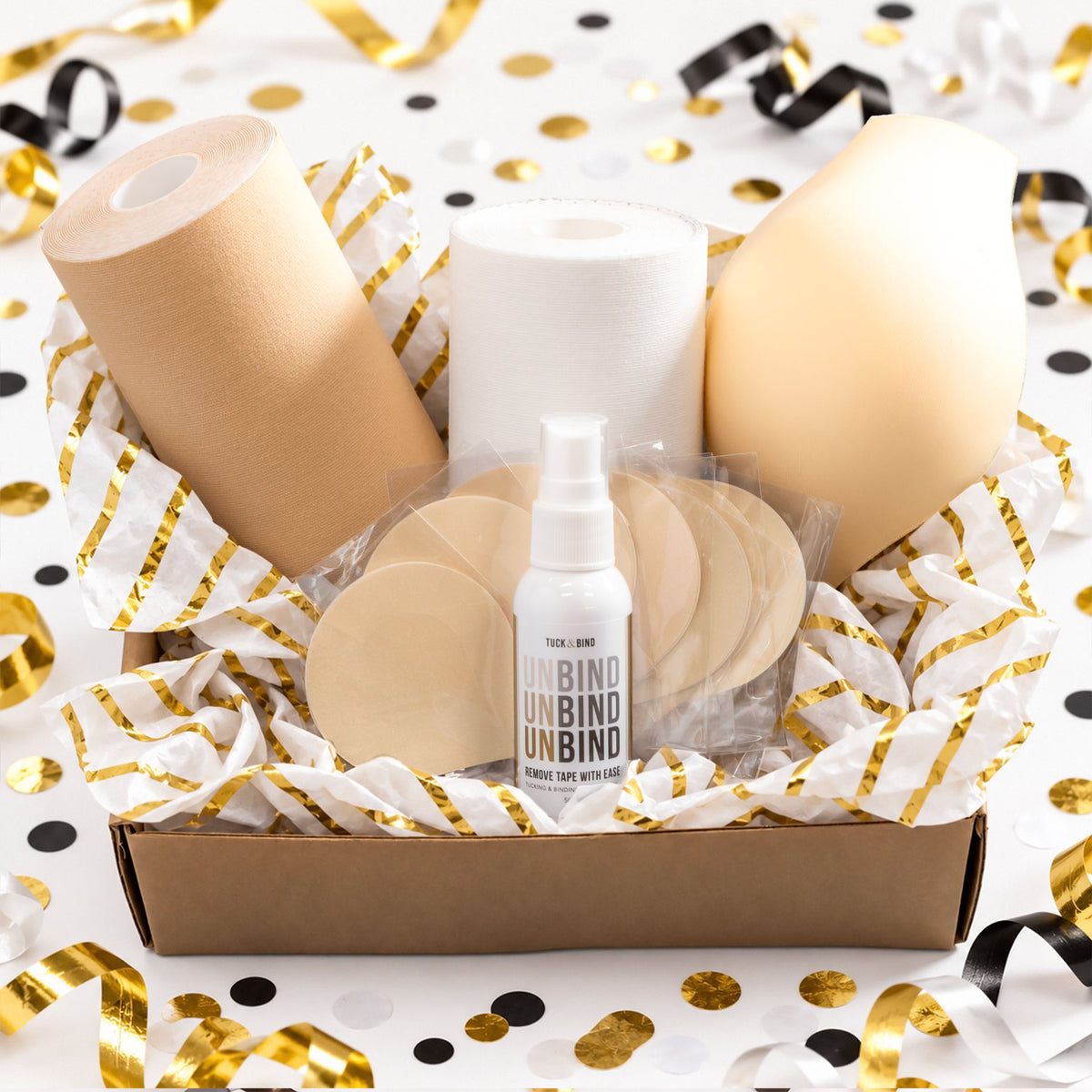 A gift box of trans masc accessories including trans tape, removal spray, nipple covers and a packer displayed on a white background with gold and black confetti.