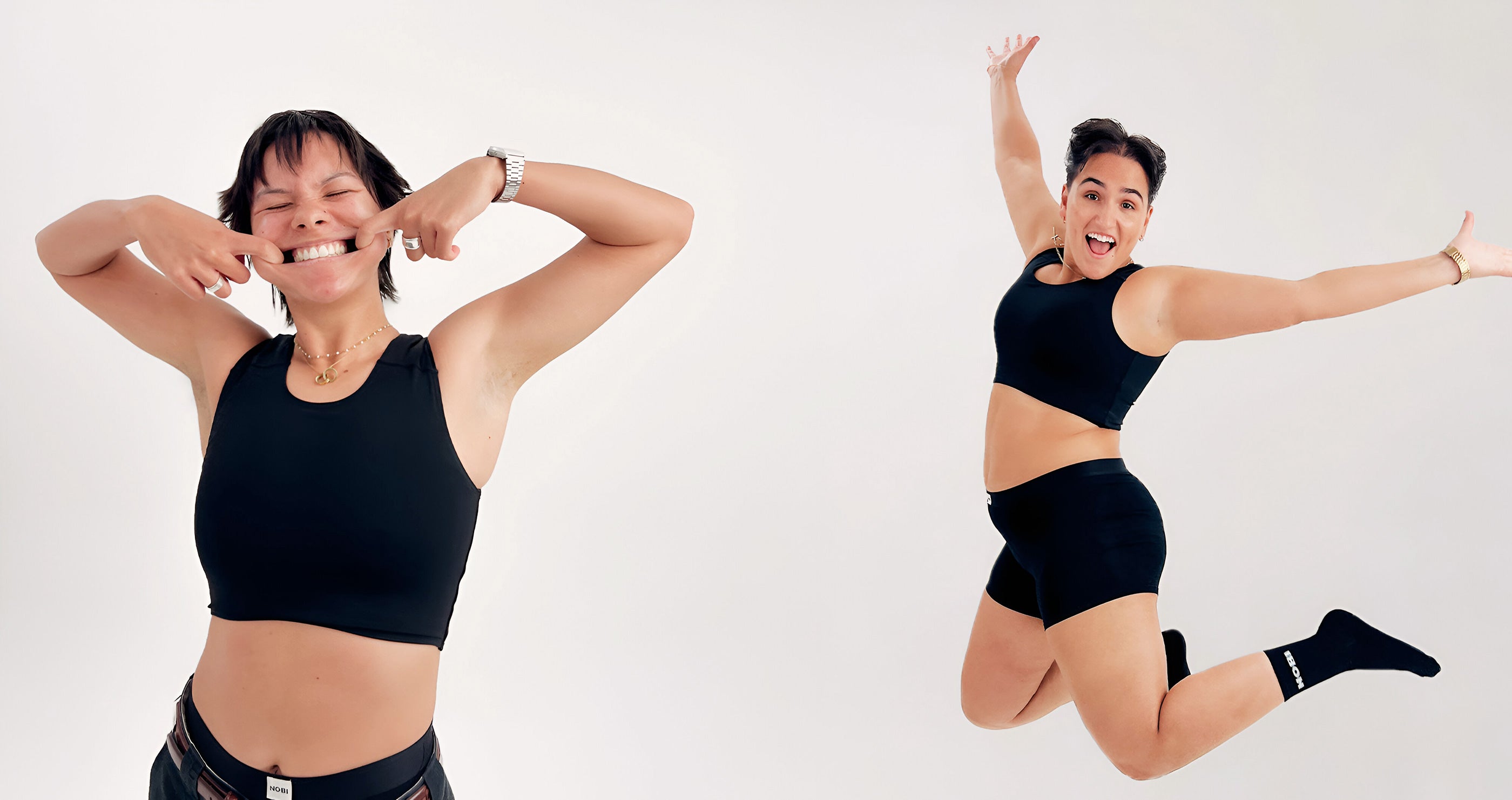 Two people wearing black chest binders. The person on the left is smiling with teeth and fingers in their mouth. The person on the right is jumping in the air with hands out while smiling.