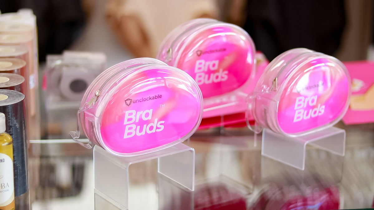 Three pink round containers labeled 'Bra Buds' and 'Unclockable' displayed on clear acrylic stands on a reflective surface, with a softly blurred indoor background.
